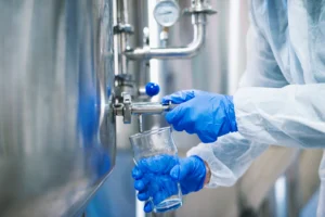 close up view hands protective gloves opening tap valve filling glass with samples from industrial machine reservoir